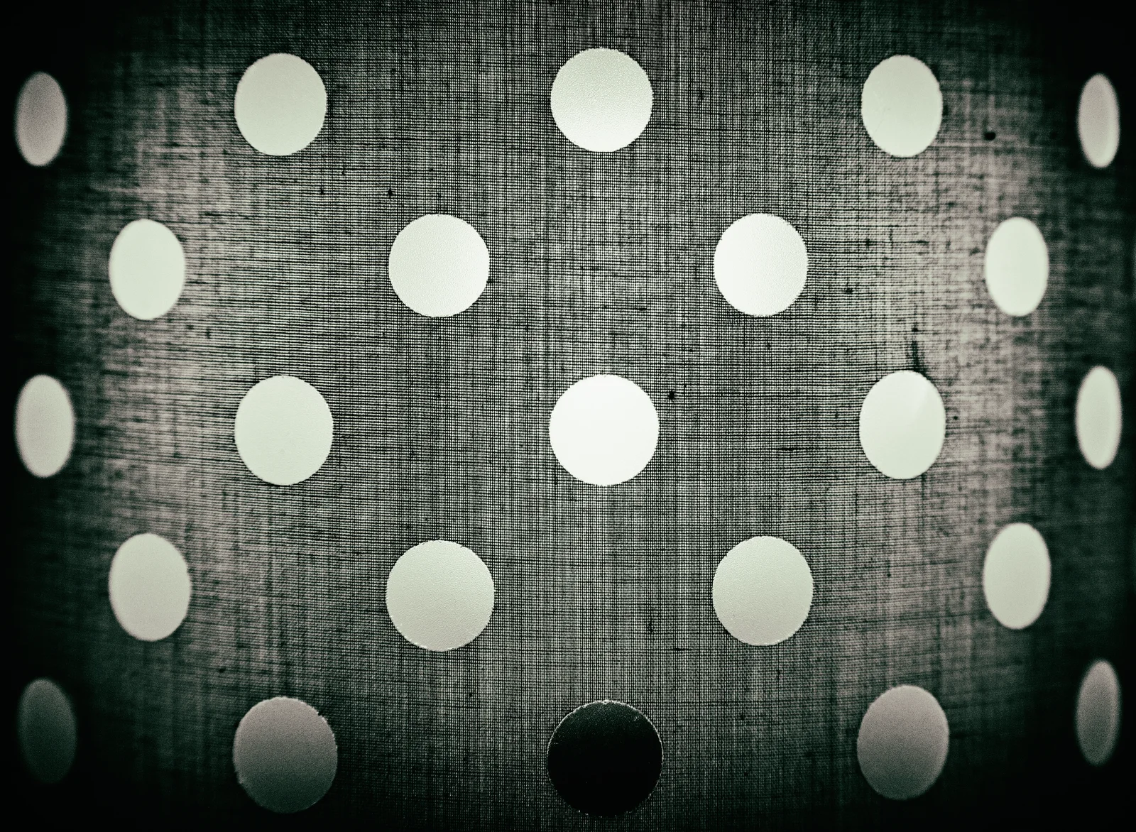 Light passes through a perforated surface, with one dark opening interrupting the pattern.