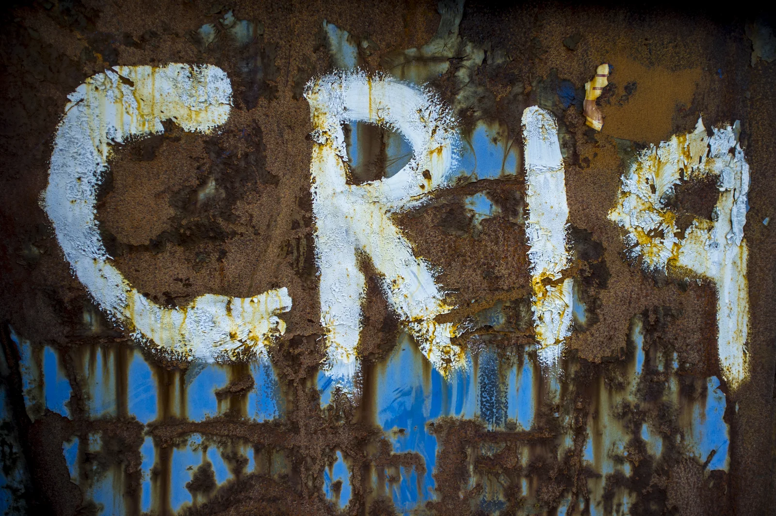 Weathered metal carries broken white letters and traces of blue paint.