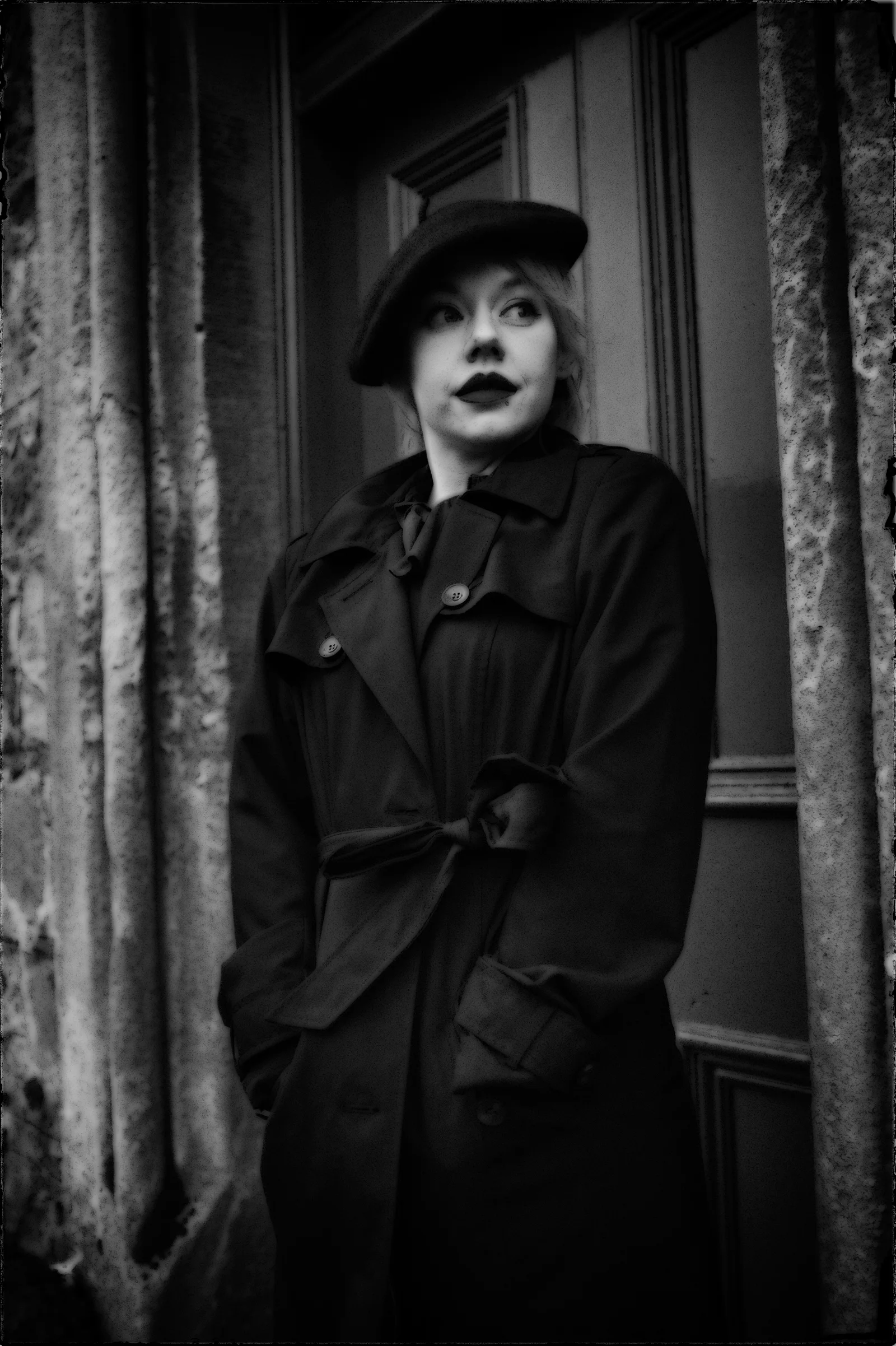 A figure in a coat and hat stands in the shadow of a doorway.