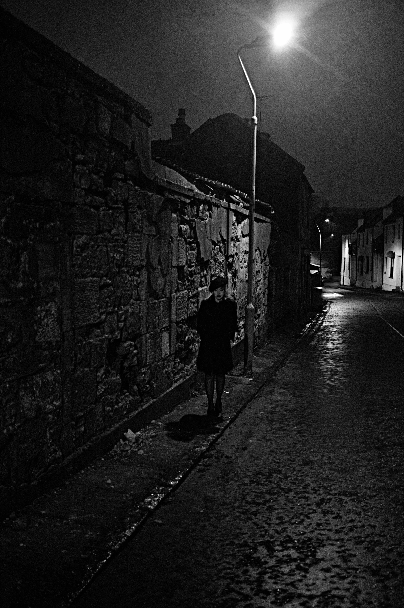 A lone figure walks down a wet monochrome street.