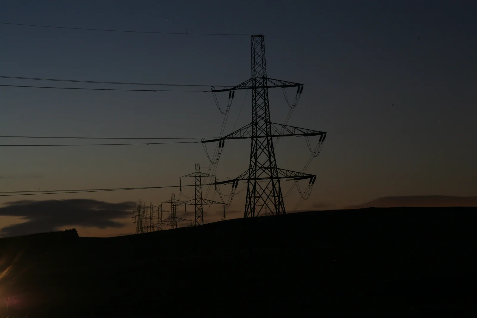 Electric pylons recede across a dark landscape at dusk.