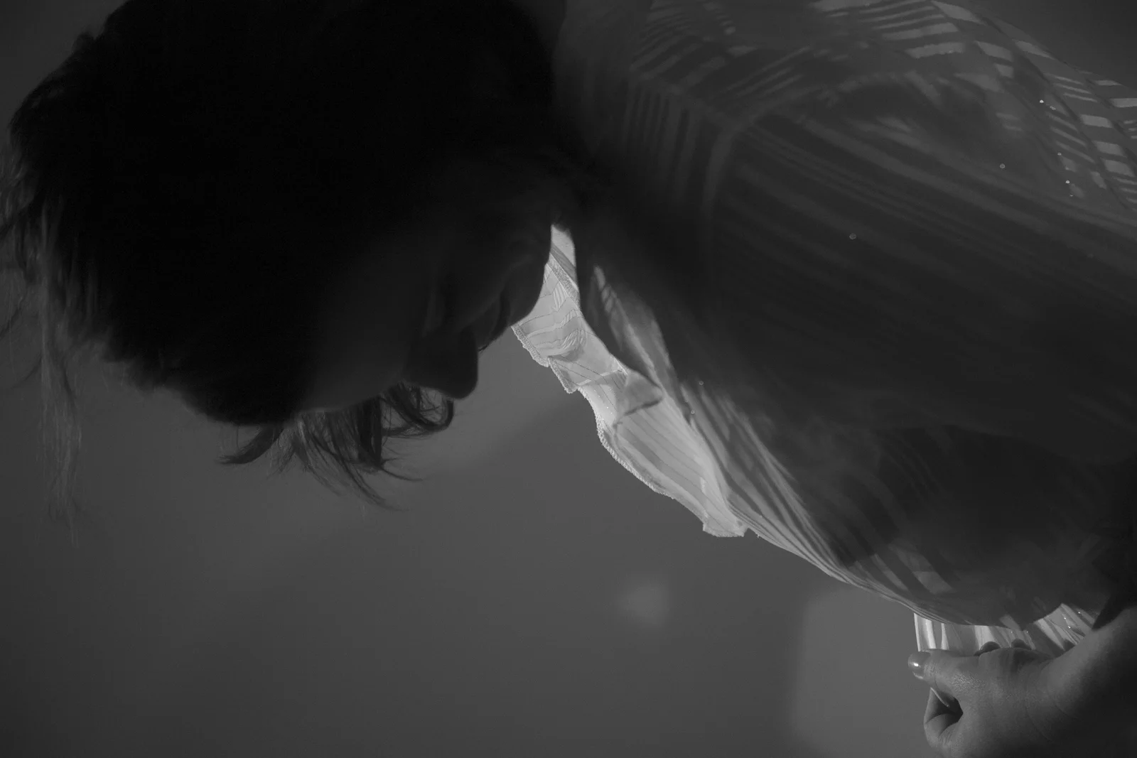 A woman bows forward as pale fabric and shadow surround her.